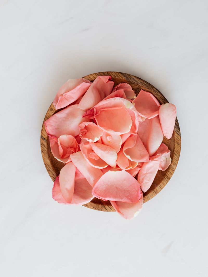 Petal Enzyme Mask — scattered rose petals in a wooden bowl on white marble surface, overhead soft natural light