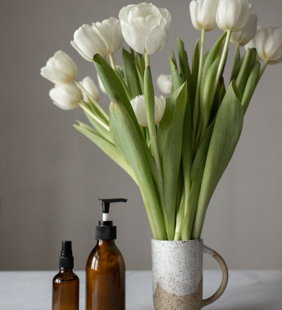 The Morning Ritual Set — white tulips in a ceramic vessel with amber glass skincare bottles on a clean neutral shelf, soft editorial light